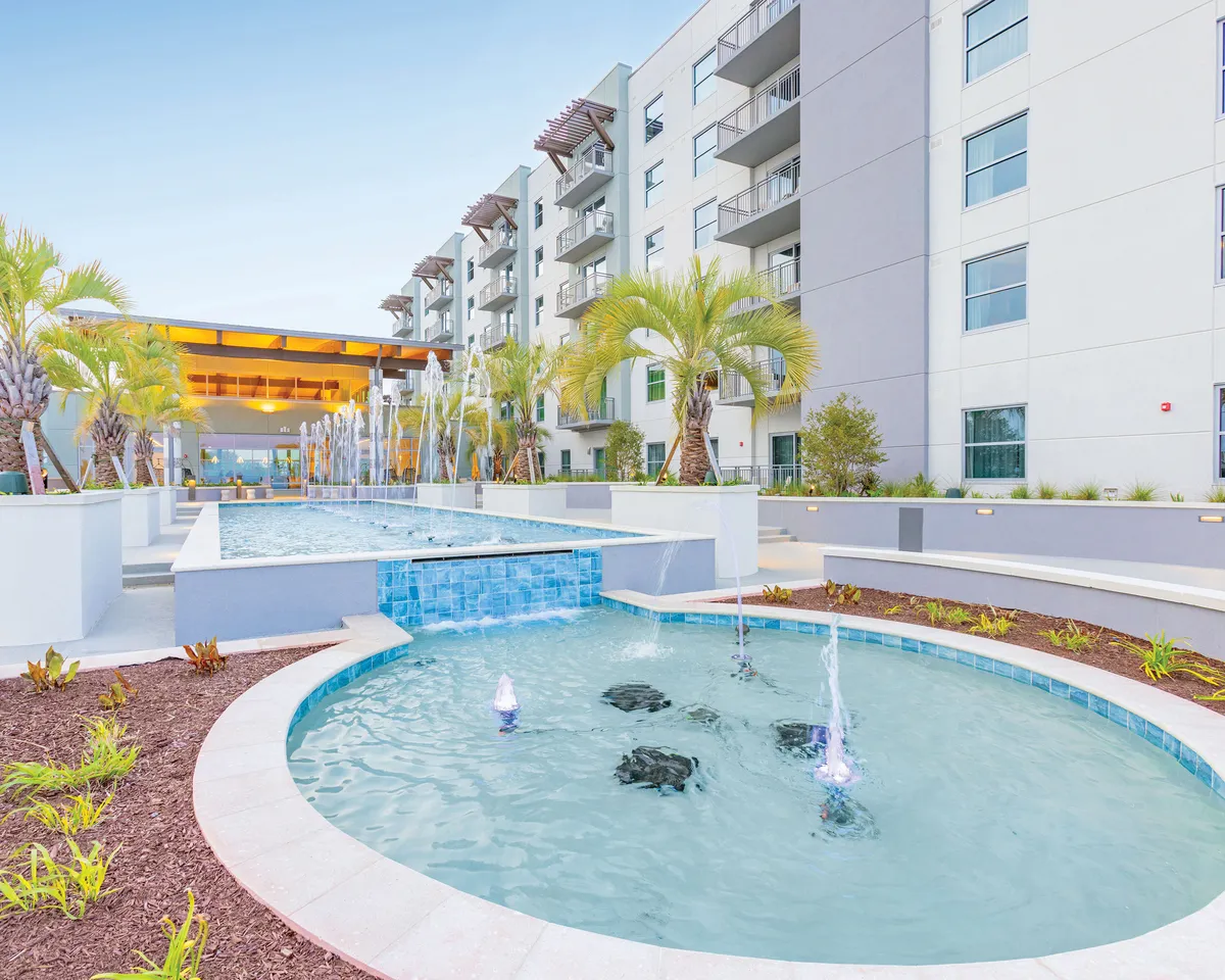 A vibrant exterior scene showing a sparkling water feature surrounded by lush palm trees, with the resort's building facade in the background. The area is designed for relaxation and social gatherings, with ample space for guests to lounge by the pool or enjoy the tranquility of the fountains. Ambient lighting hints at the space being inviting both day and night.