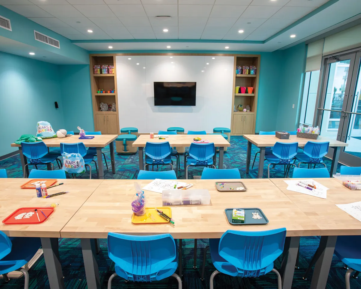 A playful, energetic space equipped with tables set for art and craft sessions, surrounded by colorful chairs. The room is filled with shelves displaying a variety of creative items and a TV screen, setting the scene for interactive group activities, workshops, or memorable gatherings.