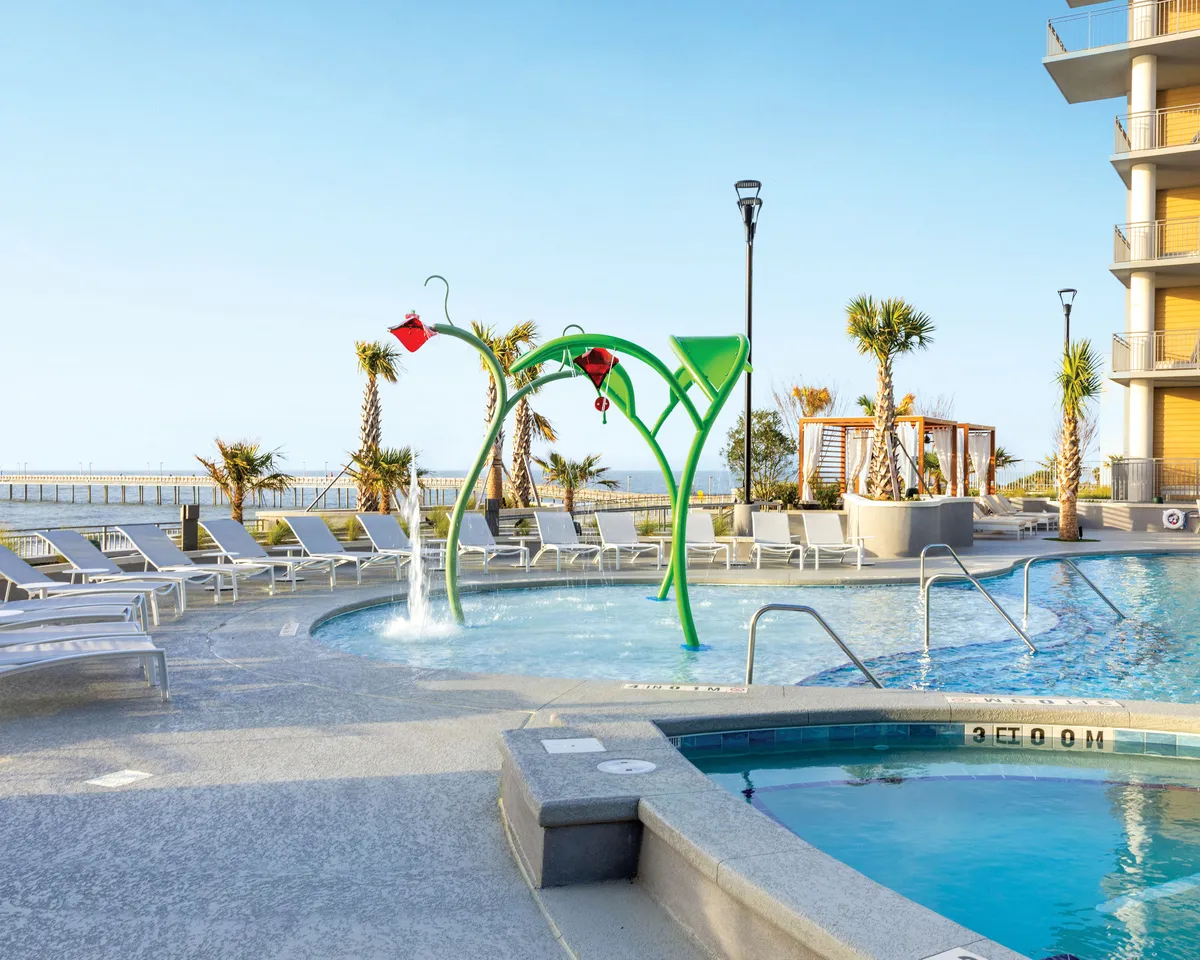 A playful and relaxing pool area with a prominent water feature where children are actively playing. Palm trees and numerous lounge chairs add tropical flair, while the setting sun hints at a pleasant evening ambiance. Families can be seen enjoying themselves, giving the scene a lively, welcoming vibe.