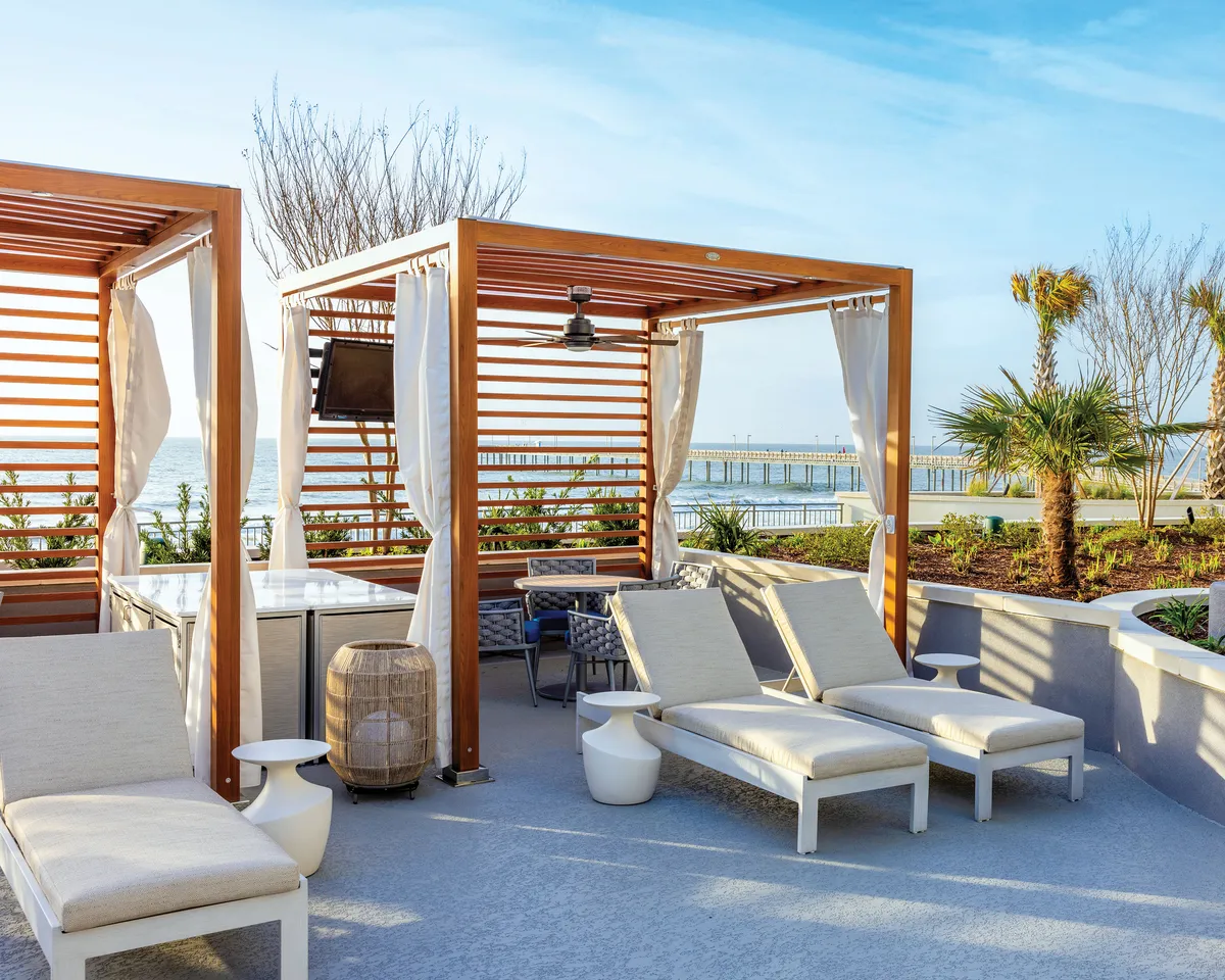 This image showcases a tranquil outdoor lounge area with wooden cabanas and lounge chairs facing a stunning ocean view. Palm trees frame the space, and the ambiance suggests a serene, private escape perfect for relaxation while soaking in the oceanfront setting.