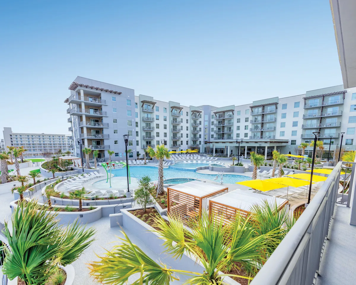 A contemporary, expansive pool surrounded by modern apartment buildings and lush, landscaped gardens with palm trees. Sun loungers under bright yellow umbrellas create inviting spaces for relaxation, while the large pool invites both swimming and socializing. The overall atmosphere is relaxed and cheerful, perfect for family gatherings or sunset swims.