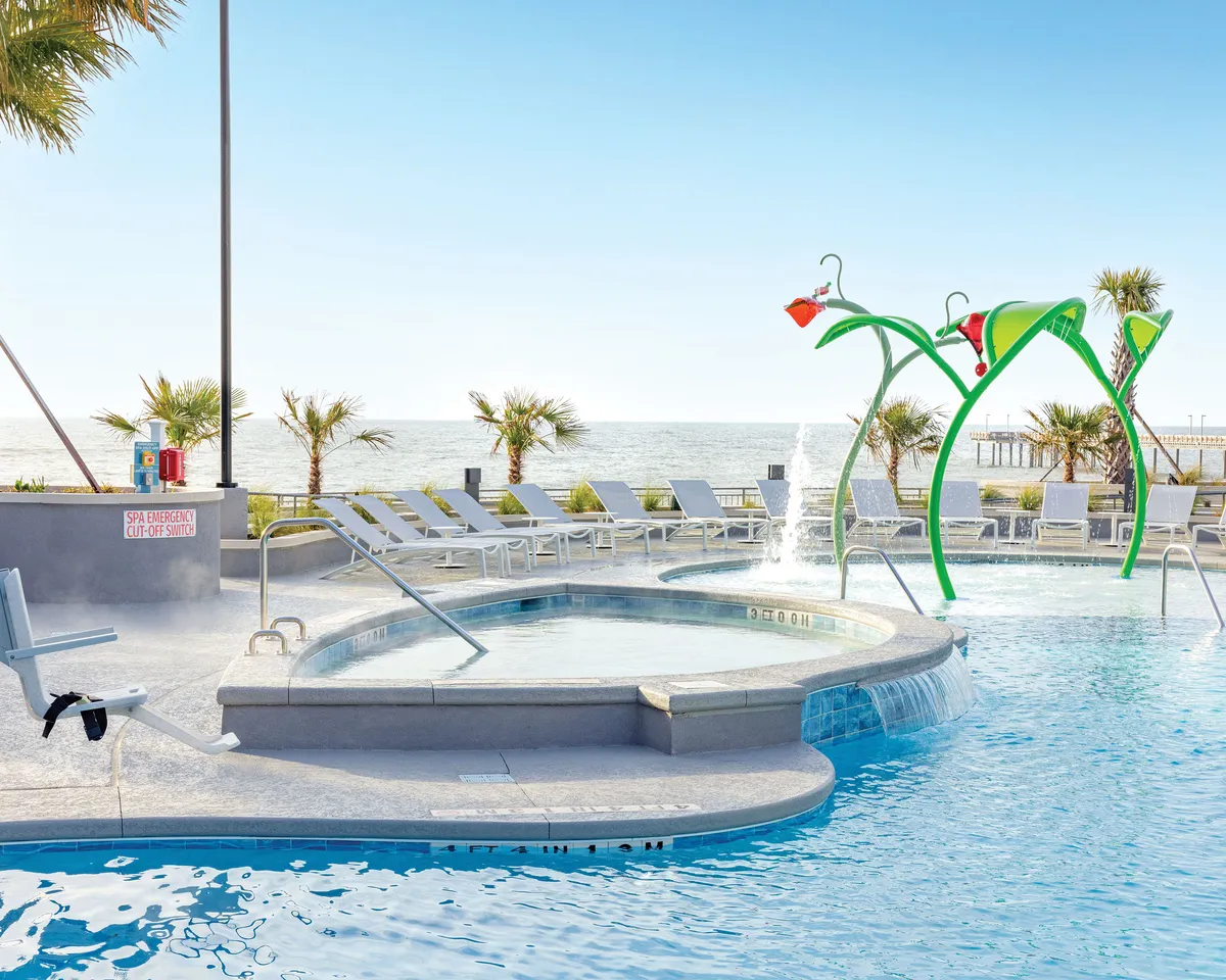 A vibrant exterior scene featuring a swimming pool with a colorful water play structure, surrounded by lounge chairs and an inviting ocean view in the background. Families are seen playing in the pool, while others sunbathe and relax by the water's edge.
