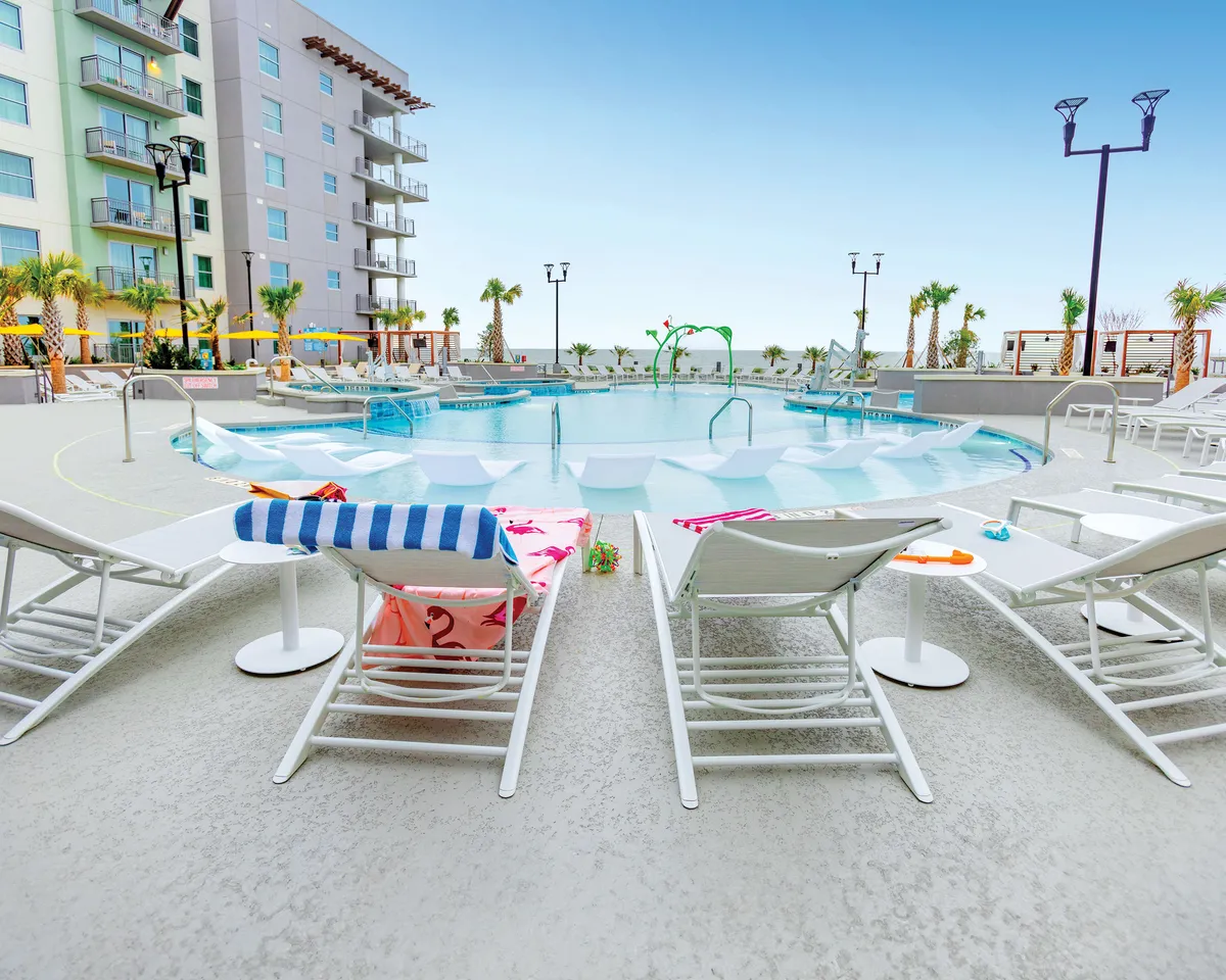 A vibrant pool scene featuring the resort's swimming pool surrounded by lounge chairs and towering palm trees, with the building's facade in the background. The area is lively, with sunbathers, swimmers, and families relaxing or socializing. The sunset in the sky casts a cheerful, golden glow over the water and pool deck, evoking a sense of fun and relaxation.