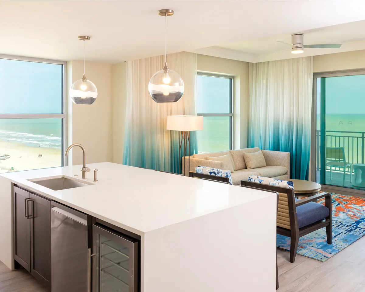 A spacious open-concept living area featuring a kitchen island with upgraded appliances, a comfortable sofa set atop a decorative rug, and large windows offering stunning ocean views. The space is filled with natural light and is perfect for both cooking and relaxing while enjoying the scenic surroundings.