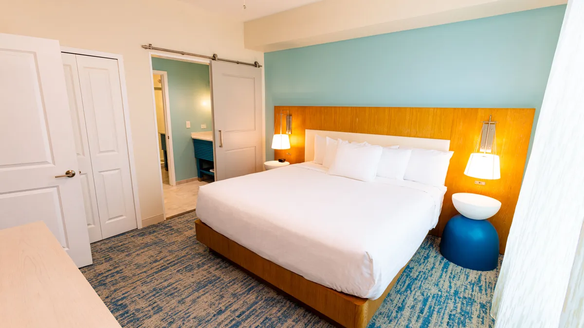 An interior scene featuring a king-sized bed, an accent wall, a blue accent chair, and a nightstand with a lamp. Ample natural light floods the space, creating a relaxing atmosphere ideal for mornings or evenings indoors, with potential access to the balcony visible through the bright windows.