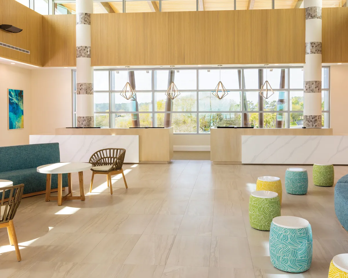 A bright, sunlit lobby area with expansive windows flooding the space with natural light. Colorful seating stools are arranged for casual conversation, and a modern reception desk stands as a central focal point. Guests are seen relaxing and socializing, creating an inviting, communal atmosphere. Art decor and stylish finishes hint at a vibrant, contemporary resort experience.